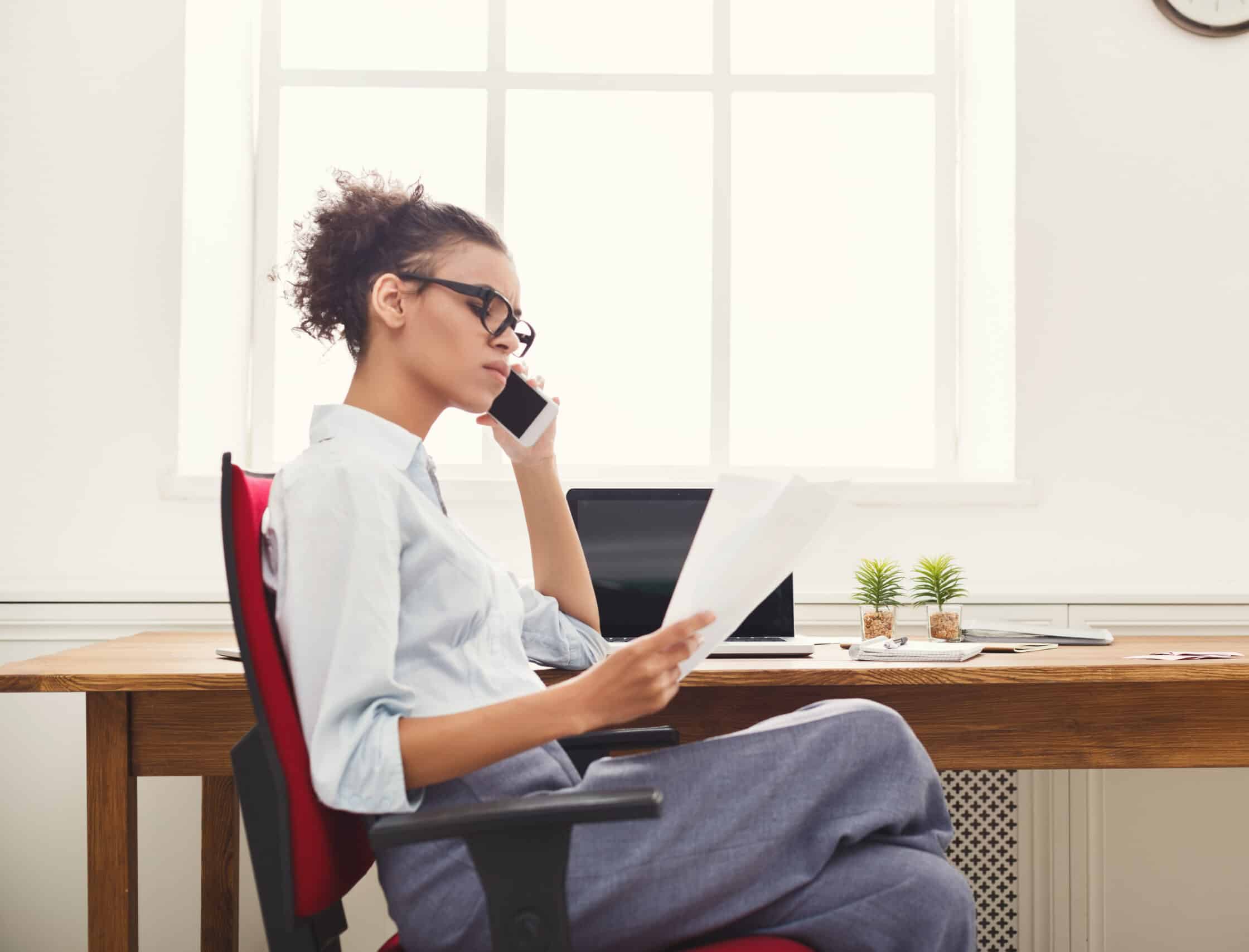 Young serious african-american businesswoman talking by phone with papers, sitting at modern office workplace, side view. Business consulting, copy space