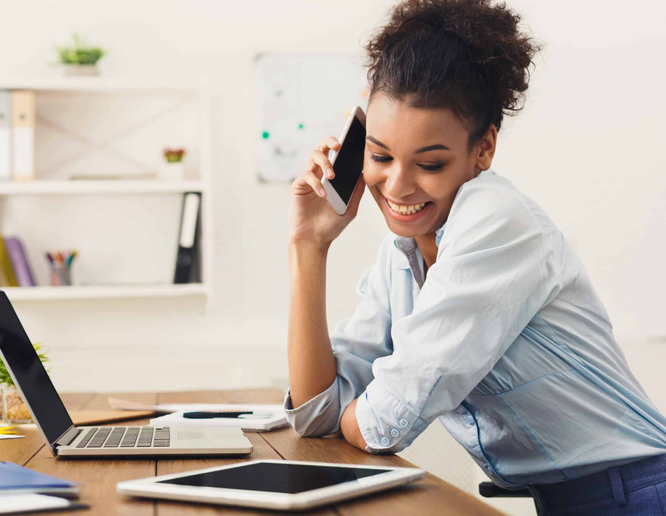 Happy african-american business woman talking on mobile at office. Young female manager consulting on phone, using laptop at working place, copy space