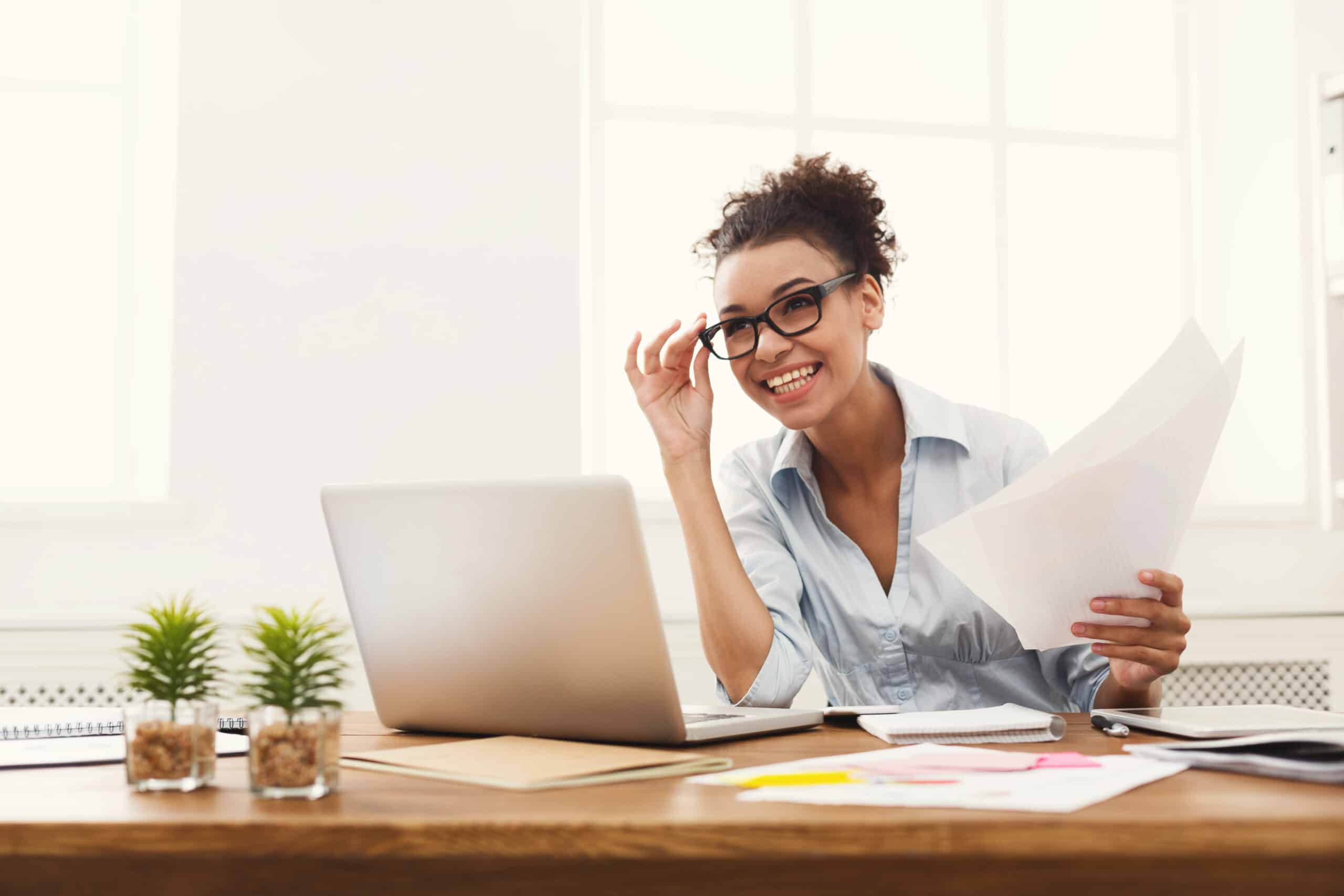 Paperwork. Happy smiling african-american business woman in formal wear sitting at wooden desk in modern office and reading report document.