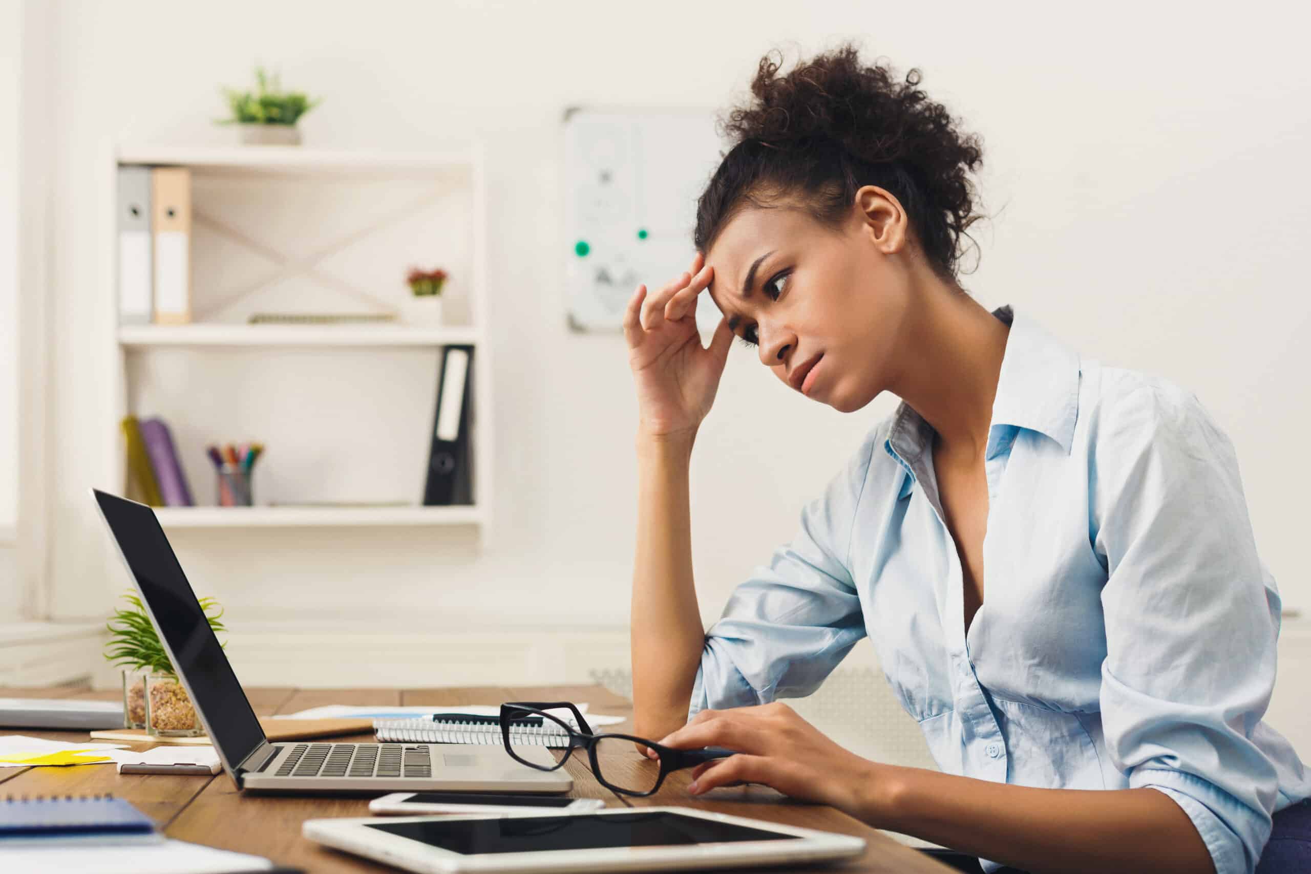 Deadline stress concept - sad african-american business woman sitting at desktop in office, working on laptop and holding hand on head. Hard working day, copy space