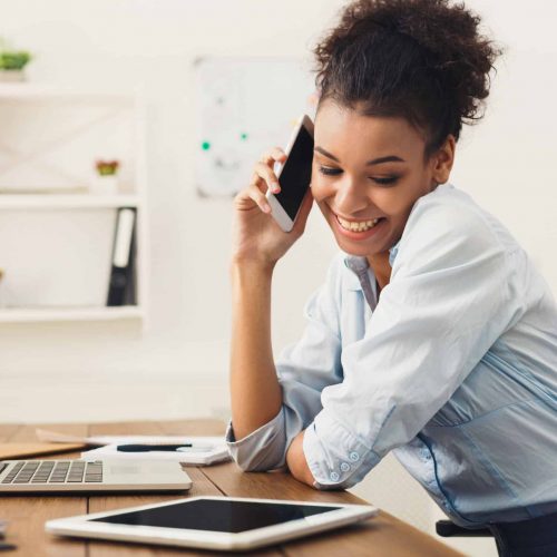 Business talk, woman consulting by phone at office Happy african-american business woman talking on mobile at office. Young female manager consulting on phone, using laptop at working place, copy space